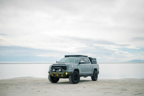 A gray off-road pickup truck with large tires and roof rack is parked on a sandy beach near a calm, expansive body of water under a cloudy sky. by Justin Salem Meyer