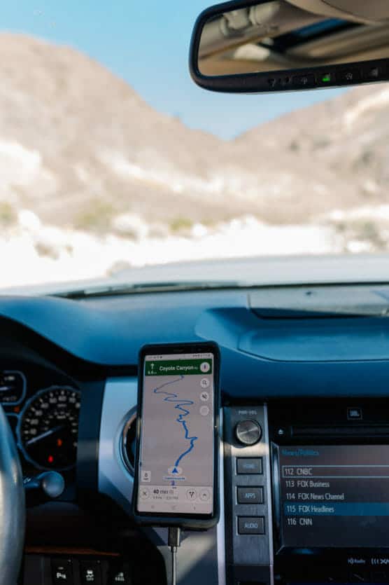 A smartphone mounted in a car displays a GPS map for Coyote Canyon. The car is parked in a desert area with rocky hills visible through the windshield. The car’s dashboard and infotainment screen are also visible. by Justin Salem Meyer