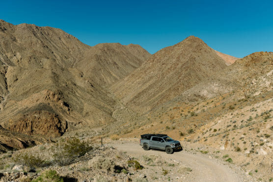 A gray off-road vehicle drives along a winding dirt road in a rugged, rocky desert landscape with brown mountains under a clear blue sky. by Justin Salem Meyer