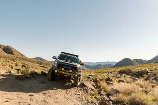 A black off-road Toyota truck with large tires drives over rocky terrain in a desert landscape with mountains in the background under a clear blue sky. by Justin Salem Meyer