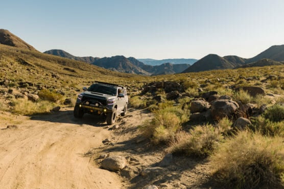 A white off-road vehicle drives along a rugged dirt trail through a rocky, mountainous desert landscape under a clear sky. by Justin Salem Meyer