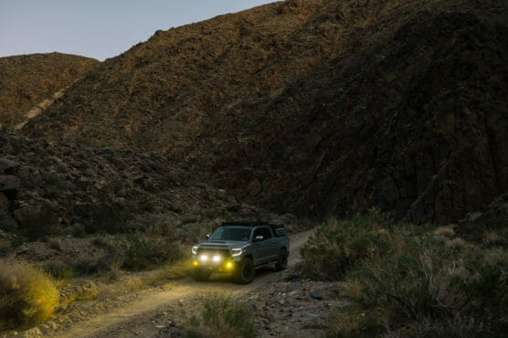A gray SUV with yellow headlights drives on a dirt road through a rocky desert landscape at dusk, surrounded by shrubs and rugged hills. by Justin Salem Meyer