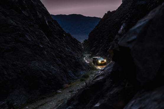 An SUV with headlights on drives through a narrow, rocky mountain pass at dusk, surrounded by dark cliffs with a faintly lit sky in the background. by Justin Salem Meyer