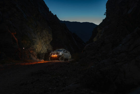 A white off-road vehicle with headlights and orange fog lights illuminates a narrow rocky canyon at dusk, with mountains silhouetted against the darkening blue sky in the background. by Justin Salem Meyer