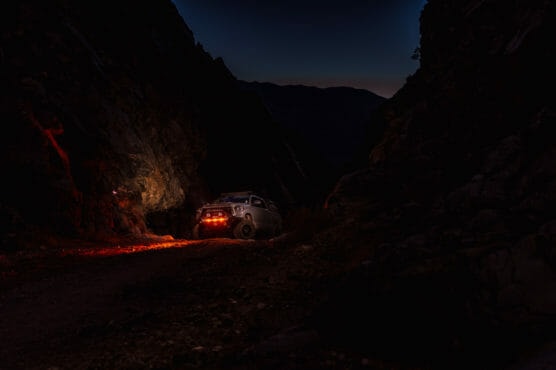 A white SUV with bright red taillights is parked on a rocky dirt road between dark mountain cliffs at night, illuminating the surrounding rocks with a red glow. The sky is dark with a faint hint of twilight. by Justin Salem Meyer