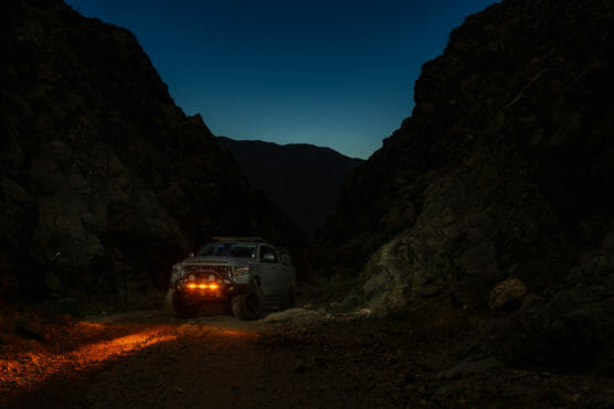 A white off-road vehicle with bright orange lights is parked on a rocky dirt trail between dark mountain cliffs at dusk or night. The headlights illuminate the ground in front of the vehicle. by Justin Salem Meyer