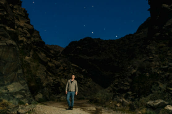 A person stands on a rocky path at night, surrounded by dark cliffs and illuminated by moonlight, with a clear sky filled with bright stars above. by Justin Salem Meyer