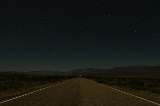 A straight, empty road stretches into the distance under a clear, star-filled night sky, with mountains faintly visible on the horizon and desert landscape on either side of the road. by Justin Salem Meyer