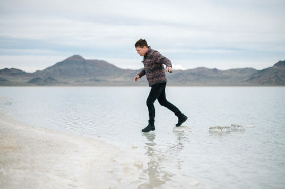 A person in a patterned sweater and black pants steps on rocks across shallow water, with mountains and a cloudy sky in the background. by Justin Salem Meyer