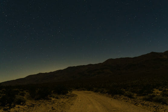 A dirt road curves through a desert landscape at night, with dark mountains in the background and a clear sky filled with bright stars overhead. by Justin Salem Meyer