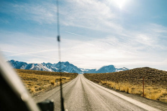 A paved road stretches into the distance through dry grasslands, leading toward snow-capped mountains under a bright blue sky with scattered clouds and a visible contrail. The photo is taken from inside a vehicle. by Justin Salem Meyer