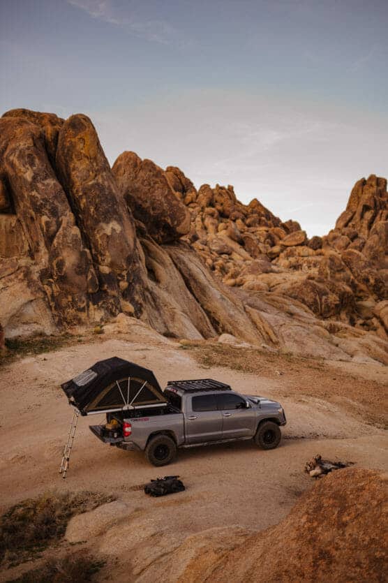 A gray pickup truck with an open rooftop tent is parked on sandy ground surrounded by large rocky formations in a desert landscape at sunset. Camping gear is set up nearby on the ground. by Justin Salem Meyer