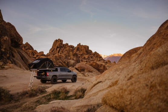 A gray pickup truck with a rooftop tent and ladder is parked among large brown boulders in a rocky desert landscape, with mountains visible in the background under a clear sky. by Justin Salem Meyer