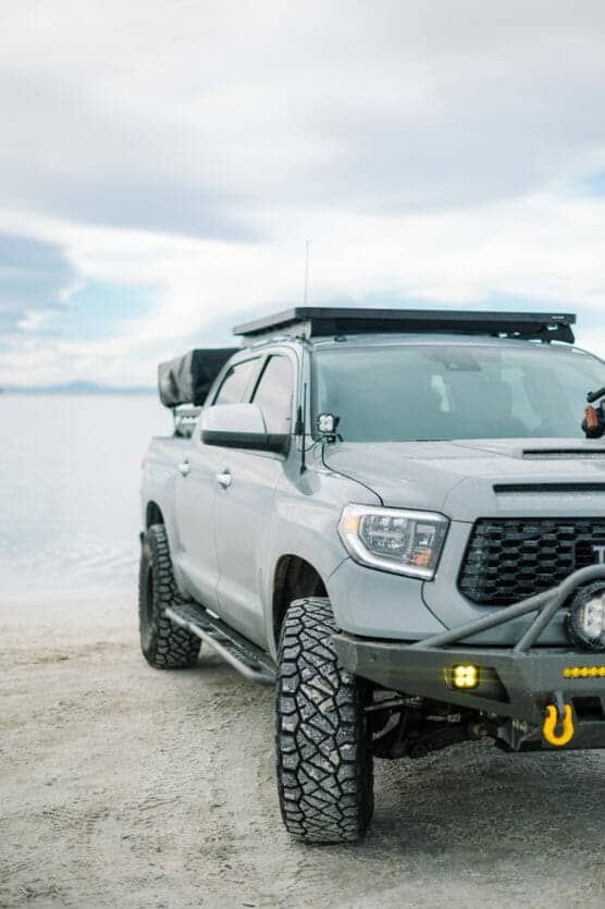 A rugged grey pickup truck with off-road tires and roof rack is parked on a sandy surface near water, under a cloudy sky. Only the front half of the truck is visible. by Justin Salem Meyer