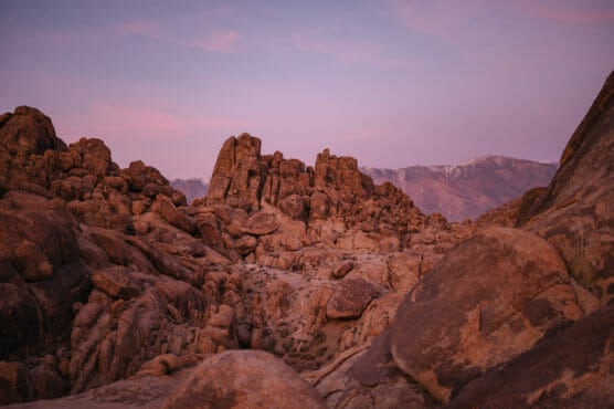 Rocky desert landscape with large reddish-brown boulders and rugged formations under a purple and pink sky at sunset; distant mountains visible in the background. by Justin Salem Meyer