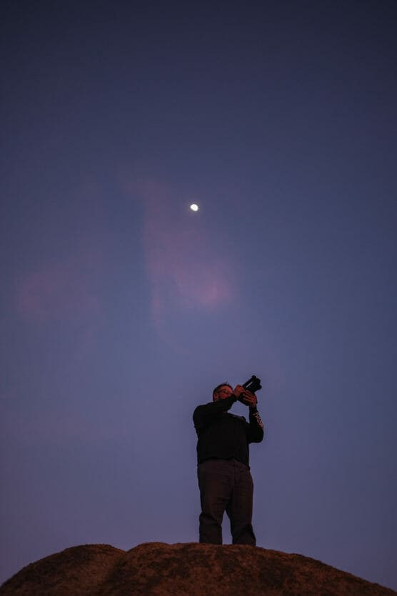 A person stands on a rocky surface at dusk, holding up a camera toward the sky. The moon is visible above in a clear, deep blue sky with faint pink clouds. by Justin Salem Meyer