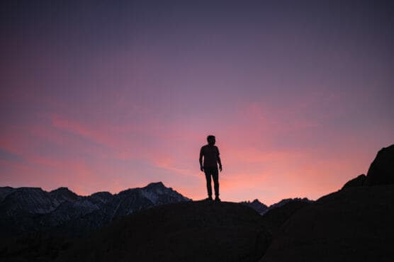 A person stands on a rocky hill silhouetted against a colorful sunset sky, with mountain peaks visible in the background. by Justin Salem Meyer