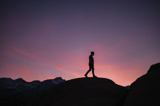 A person is silhouetted against a colorful sunset sky, walking on rocky terrain with mountain peaks visible in the background. by Justin Salem Meyer