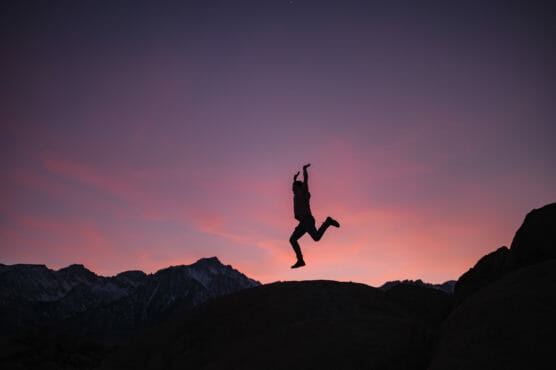 A person is captured mid-jump in silhouette against a colorful sunset sky, with mountain peaks and rocky terrain visible in the background. by Justin Salem Meyer