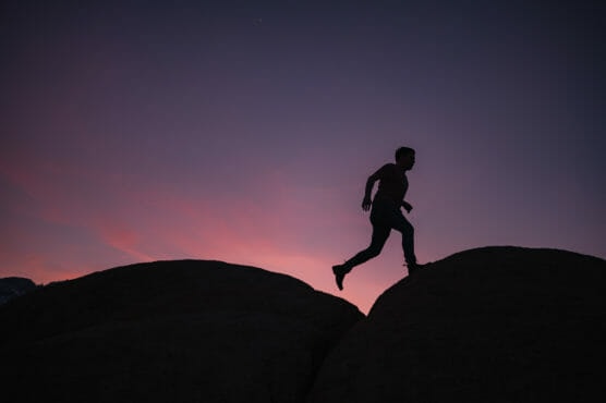Silhouette of a person running or jumping over large rocks at dusk, with a colorful sunset sky in the background. by Justin Salem Meyer