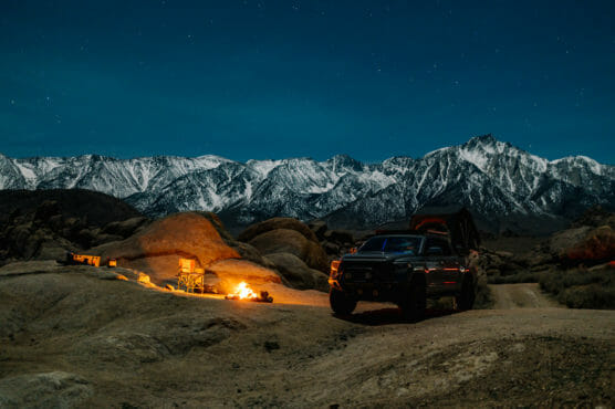 A campsite at night features a campfire, chairs, a tent, and a truck parked nearby, all set against a backdrop of snow-capped mountains under a starry sky. by Justin Salem Meyer