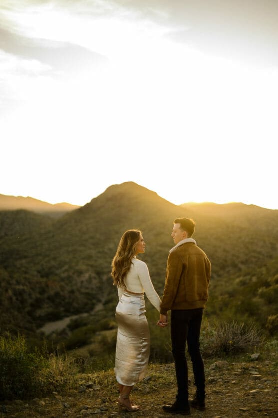 A couple stands holding hands on a hillside at sunset, facing each other with mountains and a glowing sky in the background. The woman wears a white top and satin skirt; the man wears a brown jacket and black pants. by Justin Salem Meyer