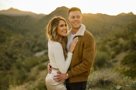 A smiling couple stands close together outdoors, with the woman’s arm around the man, against a backdrop of sunlit mountains and greenery. by Justin Salem Meyer