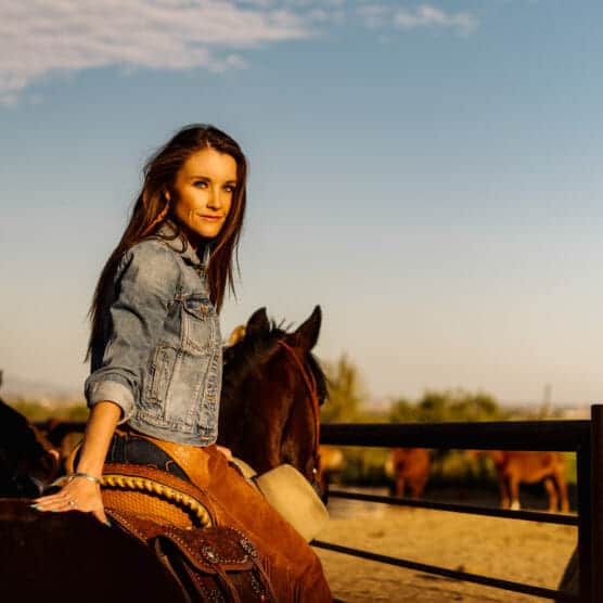 A woman wearing a denim jacket rides a brown horse in a sunlit outdoor corral, with other horses and a clear blue sky in the background. by Justin Salem Meyer