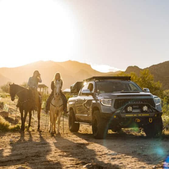 Two women ride horses next to a parked Toyota truck on a dirt path, with mountains and a bright sun in the background, creating a warm, outdoor adventure scene. by Justin Salem Meyer