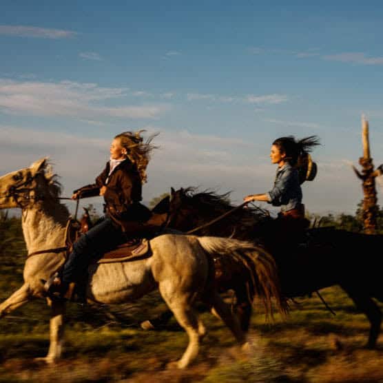 Two women ride horses at full gallop through a sunlit, grassy landscape. Their hair blows in the wind, and both wear casual jackets. The sky above is clear with a few clouds. by Justin Salem Meyer