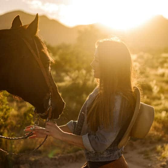 A woman wearing a denim jacket and holding a hat interacts with a horse outdoors at sunset, with sunlight streaming behind her and mountains in the background. by Justin Salem Meyer