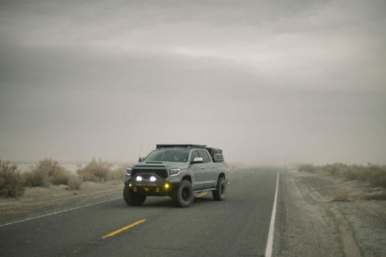A gray off-road truck with roof rack and yellow fog lights drives down an empty, foggy highway surrounded by desert landscape and sparse bushes. by Justin Salem Meyer