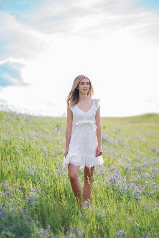 A young woman in a white dress walks through a green field with purple wildflowers under a bright, partly cloudy sky. by Justin Salem Meyer