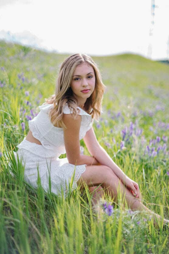 A young woman in a white eyelet dress sits in a green grassy field with purple wildflowers, looking at the camera. The sunlight is soft and the background is slightly blurred. by Justin Salem Meyer