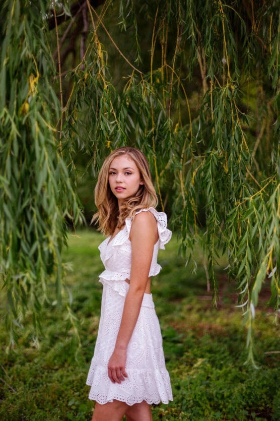 A young woman in a sleeveless white eyelet dress stands outdoors among hanging willow branches, looking at the camera with a calm expression. Lush green foliage surrounds her. by Justin Salem Meyer