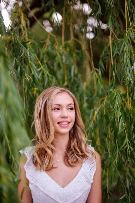 A young woman with long blonde hair and a white eyelet dress stands outdoors, smiling slightly, surrounded by green, leafy branches. by Justin Salem Meyer