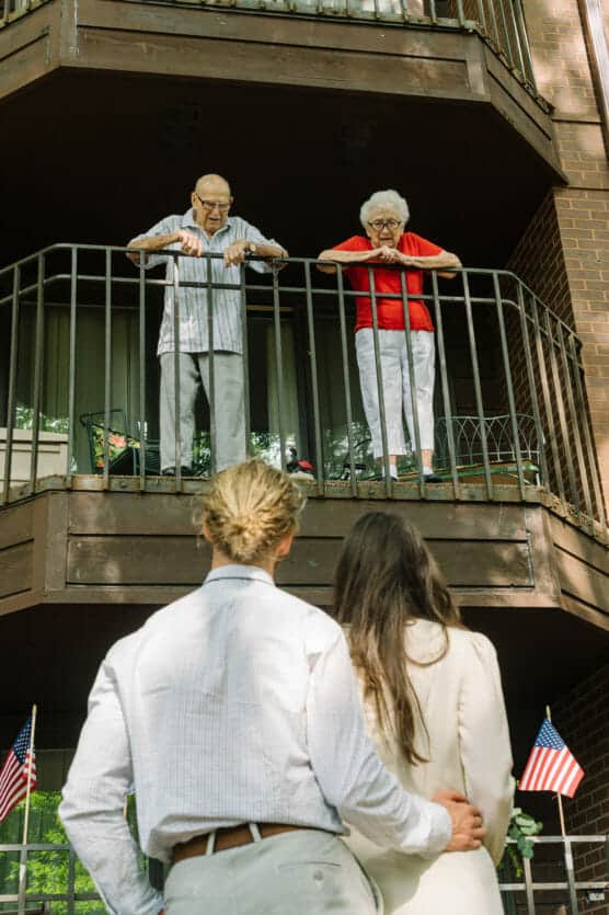 A senior man and woman stand on a balcony, looking down at a younger couple embracing below. The scene is outdoors, with small American flags visible near the bottom of the image. by Justin Salem Meyer
