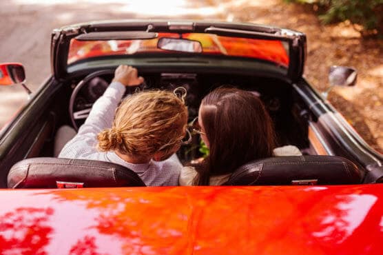 Two people sit in a red convertible car with the top down, seen from above and behind. The driver points ahead while the passenger looks in the same direction. Sunlight reflects off the car’s hood, and trees are visible nearby. by Justin Salem Meyer