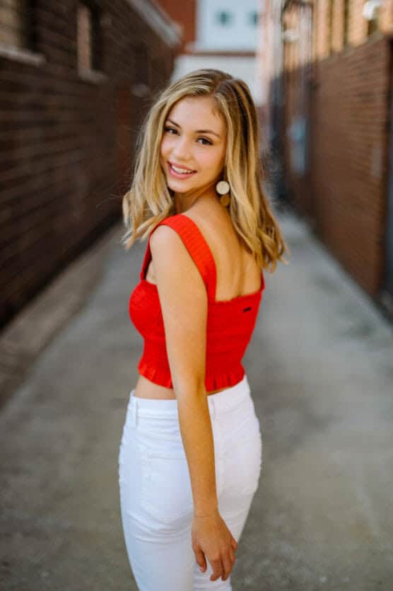 A young woman with long blonde hair, wearing a red sleeveless top and white pants, stands in an alleyway and smiles back over her shoulder at the camera. by Justin Salem Meyer