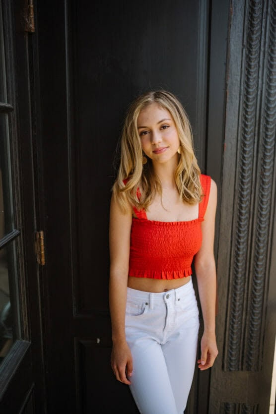 A young woman with blonde hair stands in front of a dark door, wearing a bright red sleeveless top and high-waisted white pants, smiling softly at the camera. by Justin Salem Meyer