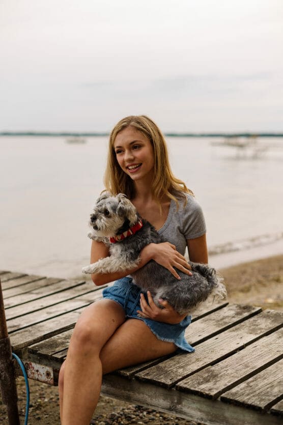 A young woman with blonde hair, wearing a gray t-shirt and denim shorts, sits on a wooden dock by the water, smiling and holding a small gray and white dog with a red collar. by Justin Salem Meyer