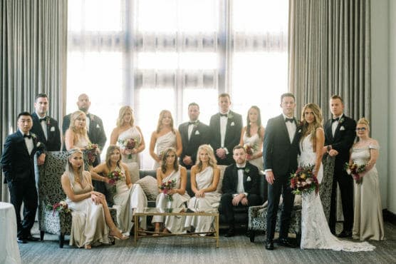 A wedding party poses indoors in formal attire. The bride and groom stand at the front, surrounded by bridesmaids in light dresses and groomsmen in dark suits. Some are seated on chairs and a couch; bouquets are held by bridesmaids. by Justin Salem Meyer