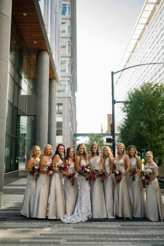 A bride in a white gown stands outdoors with nine bridesmaids in light-colored dresses, all holding bouquets, posing together in a modern city setting with tall buildings and trees. by Justin Salem Meyer