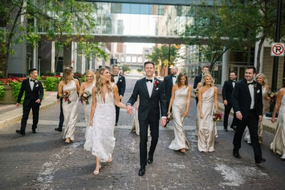 A bride and groom walk hand in hand down a city street, smiling, surrounded by bridesmaids in light dresses and groomsmen in black suits, all looking joyful. Trees and buildings line the street. by Justin Salem Meyer