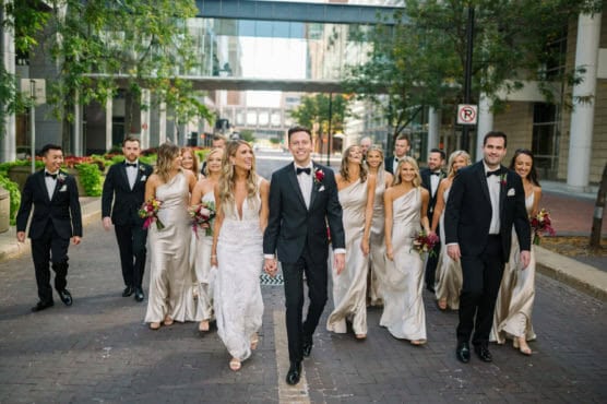 A bride and groom walk hand in hand down a city street, smiling, surrounded by their wedding party in formal wear. Bridesmaids wear cream dresses and groomsmen wear black tuxedos. Trees and modern buildings are in the background. by Justin Salem Meyer