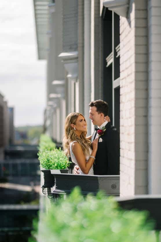 A bride and groom stand close together on a small balcony, smiling at each other. The bride holds a bouquet, and green plants decorate the railing. Buildings and a cityscape are visible in the background. by Justin Salem Meyer