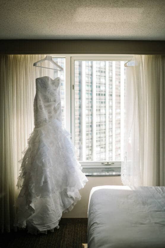 A white, ruffled wedding dress and a sheer veil hang on hangers in front of a window with city buildings visible outside. Soft light filters through the curtains, illuminating the dress and veil. by Justin Salem Meyer