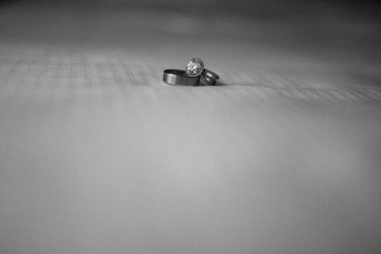 A black and white photo of two rings, one plain band and one diamond engagement ring, placed close together on a textured surface with a blurred background. by Justin Salem Meyer