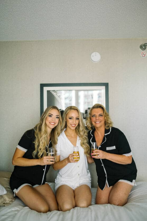 Three women sit on a bed, smiling and holding glasses of orange juice. The woman in the center wears white pajamas, while the women on either side wear matching black pajamas. They appear to be in a hotel room. by Justin Salem Meyer