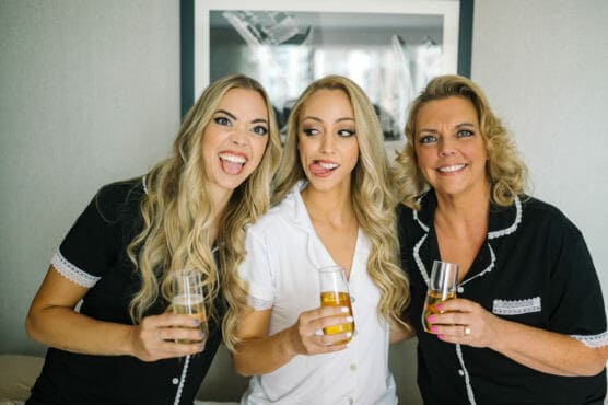 Three women with long blonde hair, wearing matching pajamas, smile and make silly faces while holding champagne glasses. They are sitting closely together in a bright room with a framed picture on the wall behind them. by Justin Salem Meyer